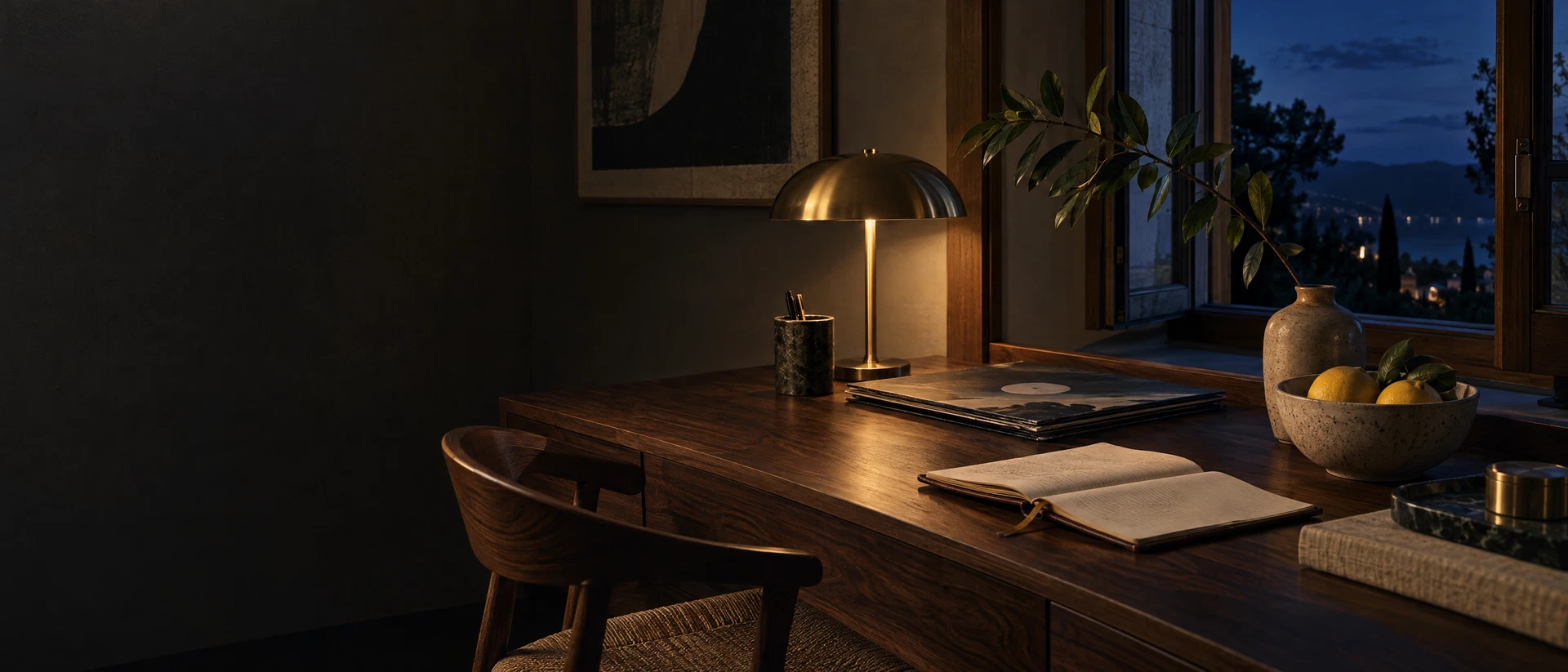 A walnut desk with a record, travel journal, ceramic lemons, and brass lamp at blue hour